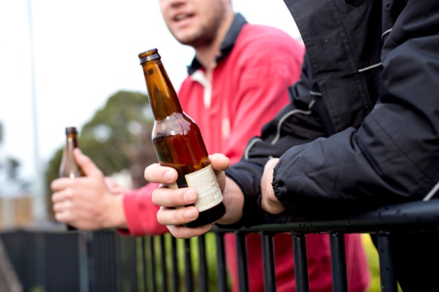 Close up of two beer bottles, each held by a different person leaning on a metal fence.