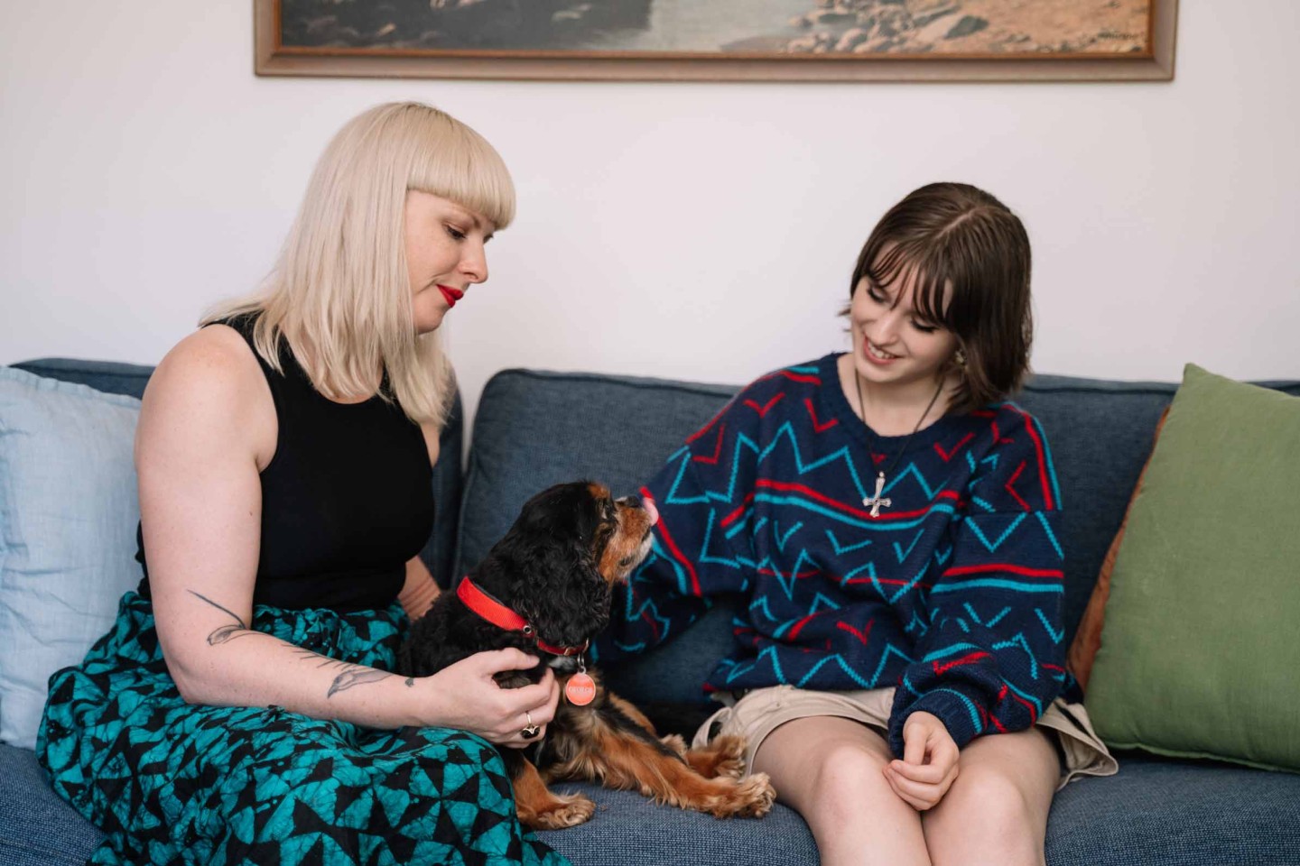 An adult and their young adult sitting on a couch, petting a dog